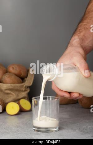 Une main verse du lait de pomme de terre crémeux d'une bouteille en verre dans un verre transparent. Les pommes de terre fraîches sont placées dans un sac en tissu à proximité, créant un kitche rustique Banque D'Images