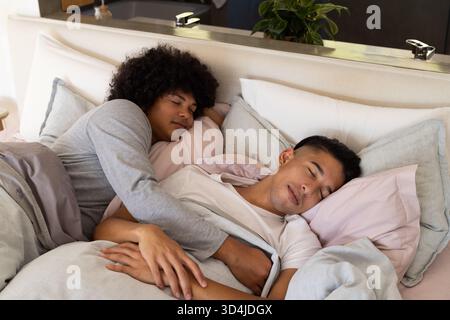 Câlin divers partenaires masculins reposant sur le lit dans la chambre, avec des oreillers roses et des plantes en pot Banque D'Images