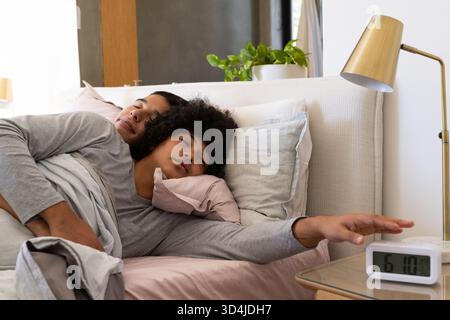 Divers partenaires masculins couchés et atteignant dans une chambre confortable, avec réveil numérique sur la table de chevet Banque D'Images