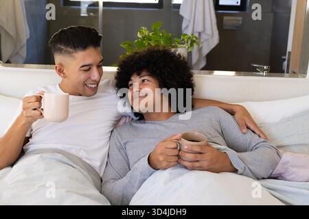 Tenant des tasses, couple masculin diversifié se penchant près dans le lit dans la chambre, avec des plantes en pot Banque D'Images