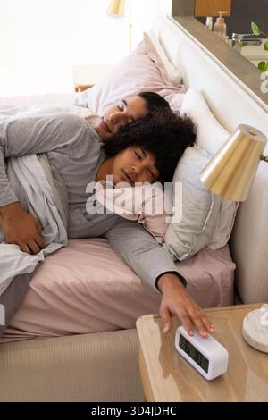 Dormir divers partenaires masculins atteignant vers le réveil sur la table de nuit dans la chambre, avec des oreillers Banque D'Images
