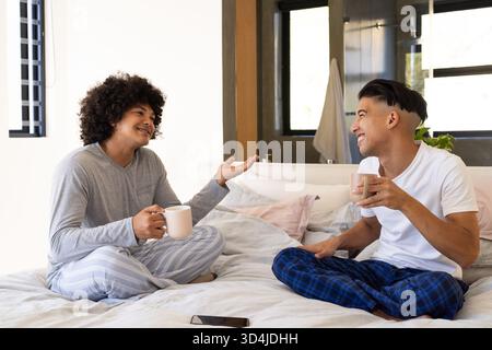 Assis divers amis masculins discutant et sirotant des tasses sur le lit dans la chambre lumineuse, avec téléphone Banque D'Images