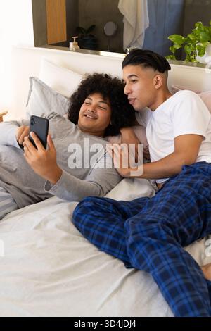 Divers amis masculins se relaxant sur le lit avec des oreillers dans la chambre, avec smartphone et plante en pot Banque D'Images