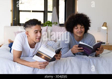 Lecture de divers amis masculins allongés sur la literie grise dans la chambre, avec livres de poche et smartphone Banque D'Images