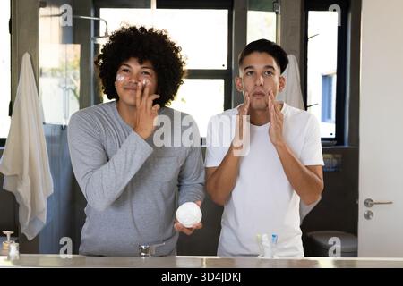 Application de crème pour le visage, divers amis masculins debout à l'évier de salle de bain, avec pot ouvert et miroir Banque D'Images