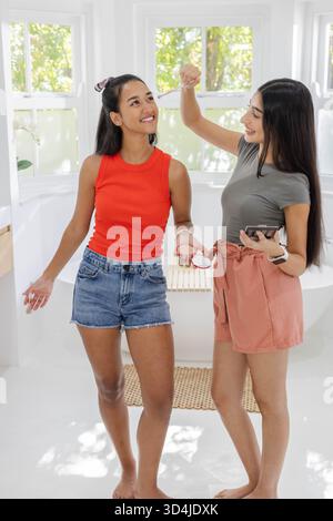 Souriant diverses amies féminines appliquant le maquillage dans la salle de bain à la maison, avec de la poudre compacte, plante d'orchidée Banque D'Images