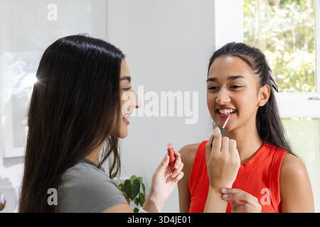 Application de brillant à lèvres diverses amies féminines debout à l'intérieur à l'espace vanité, avec la plante en pot Banque D'Images