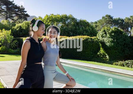 Amies féminines souriantes debout sur la terrasse au bord de la piscine, avec écouteurs sans fil et montre de sport Banque D'Images