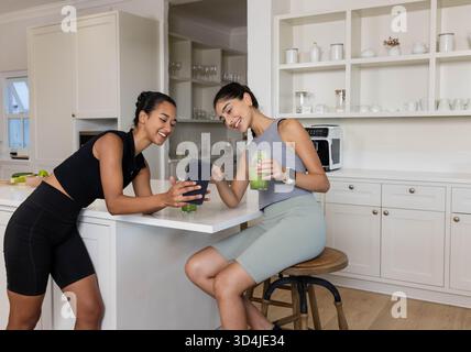 Penchant diverses amies féminines montrant smartphone sur l'îlot de cuisine blanc, avec smoothies verts Banque D'Images