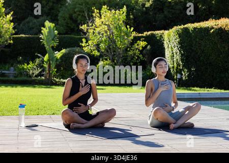 Méditer diverses amies féminines assis sur la terrasse en bois près de la piscine, avec bouteille d'eau, smartphone Banque D'Images