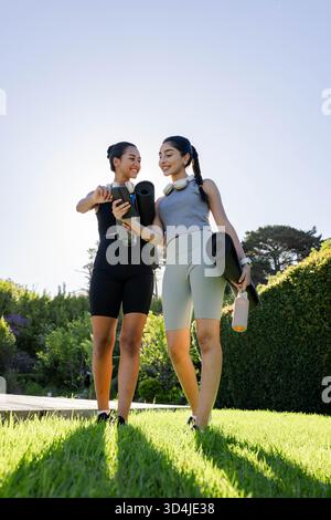 Amies féminines diverses souriantes, regardant un smartphone sur la pelouse, tenant des tapis de yoga et des bouteilles d'eau Banque D'Images