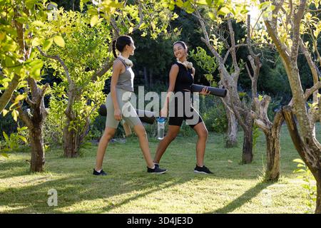 Marcher diverses amies féminines portant des tapis de yoga, des écouteurs et une bouteille d'eau à travers le jardin ensoleillé Banque D'Images