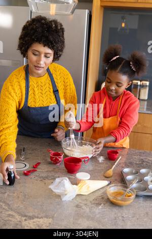 Mère et fille diverses mélangeant la pâte à l'îlot de cuisine, avec bol en verre et tasses à mesurer rouges Banque D'Images