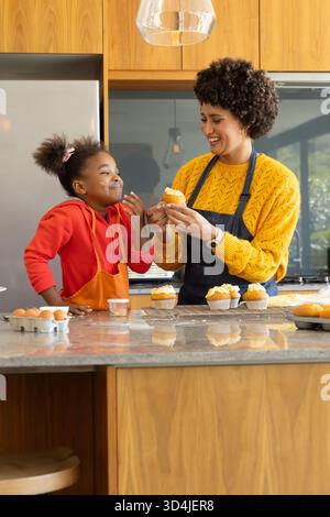 Mère et fille diversifiées décorant des cupcakes glacés dans la cuisine à la maison, avec grille de refroidissement et spatule Banque D'Images