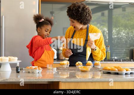 Mère et fille diversifiées décorant des cupcakes givrés dans la cuisine à la maison, avec sac à passepoil Banque D'Images