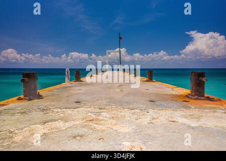 Une vue horizontale et symétrique regardant vers le bas d'une vieille jetée en béton avec des bornes rouillées, menant à l'océan turquoise vibrant sous un ciel bleu profond wi Banque D'Images