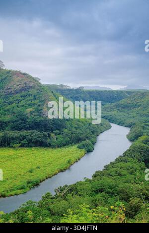 Une vue en angle élevé montre la rivière Wailua qui courbe à travers une forêt tropicale dense et verte et passe devant une prairie vert clair à Kauai, Hawaï. Le ciel est ov Banque D'Images