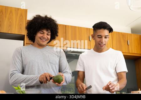 Divers amis masculins hachant des ingrédients dans la cuisine moderne, avec avocat et couteaux Banque D'Images