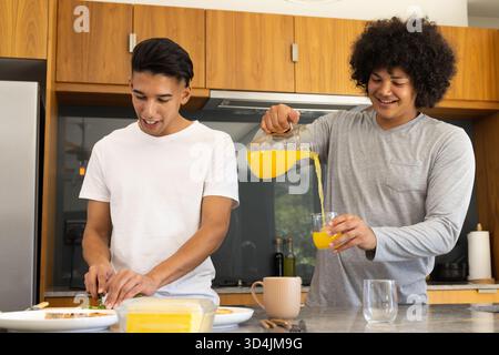 Divers amis masculins préparant le petit déjeuner dans la cuisine moderne, avec planche à découper et pichet de jus Banque D'Images
