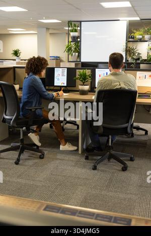 Collaboration de divers collègues examinant des graphiques à barres sur des moniteurs dans un bureau moderne, avec des usines Banque D'Images