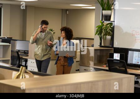 Marcher divers collègues transportant un ordinateur portable et un smartphone à travers un bureau ouvert, avec des plantes Banque D'Images