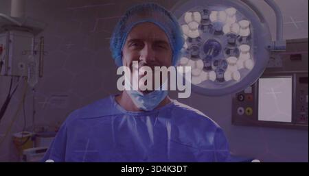 Chirurgien debout en gommages bleus, bonnet de bouffant et masque abaissé sous lumières en salle d'opération Banque D'Images