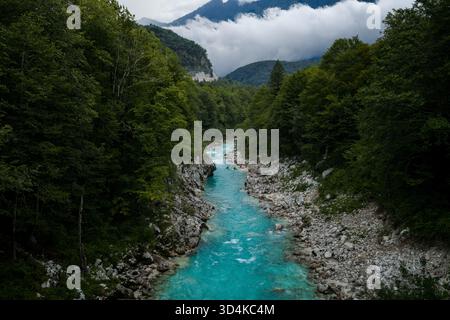 Vue aérienne de la rivière turquoise vivante Soca se fraie un chemin à travers une vallée verdoyante, embrassée par des rives rocheuses et des montagnes lointaines, embrassées par des nuages, Banque D'Images