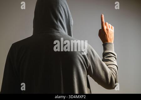 Vue arrière de l'homme en sweat à capuche levant l'index comme si vous pointez ou cliquez sur l'écran virtuel invisible, concept tech. Mise au point sélective. Banque D'Images