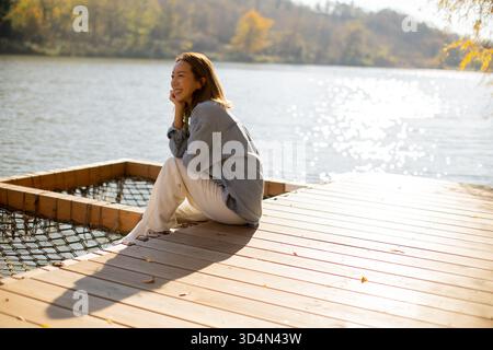 Une femme est assise paisiblement sur une terrasse en bois au bord du lac, souriante et profitant de la lumière chaude du soleil lors d'un bel après-midi d'automne. Banque D'Images