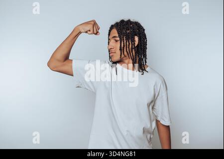 Un jeune homme se tient confiant avec son bras fléchi, montrant sa force. Il porte un simple t-shirt blanc et a des cheveux tressés, soulignant un casu Banque D'Images