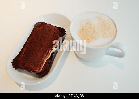 Tiramisu classique garni de poudre de cacao servi à côté d'un cappuccino mousseux sur une table blanche, cadre de café minimaliste. Le concept du petit déjeuner en A. Banque D'Images