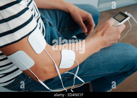 électrodes attachées au bras d'un homme lors d'une séance de thérapie d'électrostimulation à domicile, avec le dispositif électronique visible dans sa main Banque D'Images