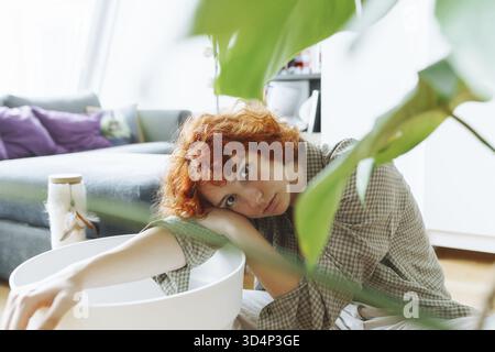 Portrait roux jeune femme attrayante engagée dans la transplantation de monstera dans un nouveau sol fertile, à la maison Banque D'Images
