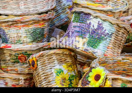 Des paniers tissés colorés avec des motifs floraux sont empilés ensemble sur un marché artisanal. La lumière du soleil met en valeur leurs textures, détails artisanaux Banque D'Images
