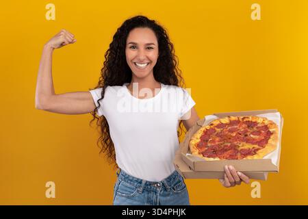 Happy Latin Woman montre de la force tout en tenant Delicious Pizza dans un studio lumineux Banque D'Images