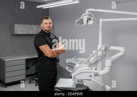 Jeune médecin dentiste masculin debout, les bras croisés, souriant à l'intérieur d'un cabinet de clinique dentaire moderne et propre Banque D'Images
