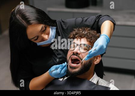 Dentiste femme portant un masque et des gants effectuant un bilan dentaire sur un patient masculin dans une clinique moderne. Se concentrer sur l'hygiène bucco-dentaire Banque D'Images