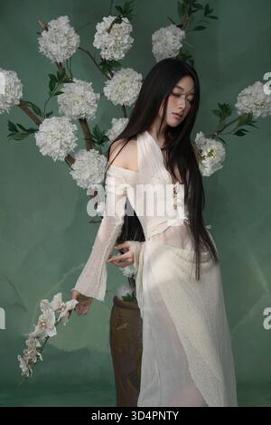 Femme asiatique éthéré dans une robe tricotée blanche avec de longs cheveux foncés, posant près d'un arbre orné de grandes fleurs blanches sur un fond vert Banque D'Images