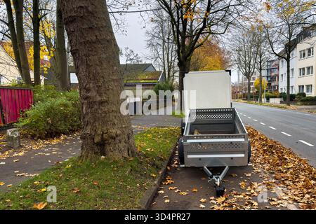 Une petite remorque est garée sur le côté d'une rue calme bordée d'arbres et de feuilles d'automne colorées. La scène présente un ciel clair, indiquant un frais Banque D'Images