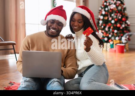 Couple aime les achats en ligne pour Noël dans le salon confortable portant des chapeaux de Père Noël Banque D'Images