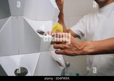 Plan recadré de jardinier mâle nettoyant le jardin vertical de la tour hydroponique avec éponge jaune dans la salle de bains moderne, assurant la propreté et optimale Banque D'Images