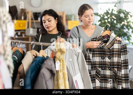 Deux jeunes femmes heureuses choisissant manteau ou veste pour la saison froide dans un magasin de vêtements avec un grand assortiment Banque D'Images