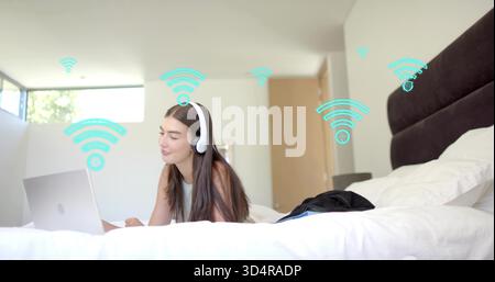 Femme couchée utilisant un ordinateur portable argenté sur le lit, portant un dessus blanc et des écouteurs, graphiques Wi-Fi sarcelle Banque D'Images
