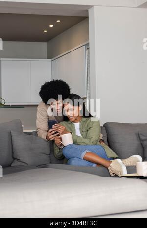 Penchant divers couple regardant smartphone sur canapé sectionnel dans le salon, tenant une tasse à café Banque D'Images