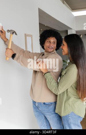 Couple diversifié installant un petit cadre en bois sur un mur blanc dans une maison moderne, avec un marteau et un clou Banque D'Images