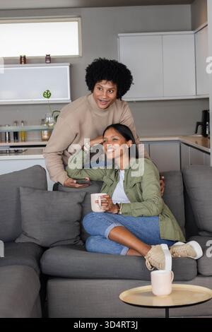 Penchant couple diversifié tenant smartphone et tasses à café sur canapé gris dans la cuisine ouverte Banque D'Images