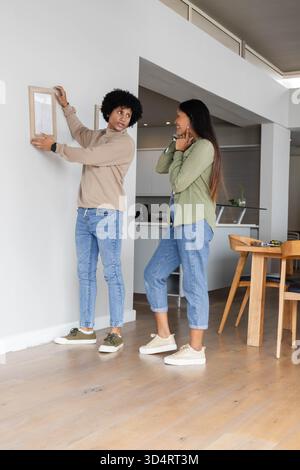 Accrocher un couple diversifié plaçant le cadre photo sur le mur dans la maison ouverte, avec mètre ruban sur la table Banque D'Images
