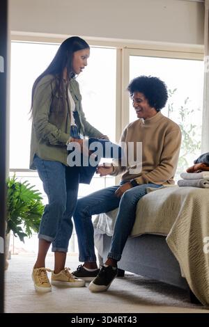 Couple diversifié remettant des vêtements pliés par lit dans une chambre ensoleillée, avec une plante verte en pot Banque D'Images