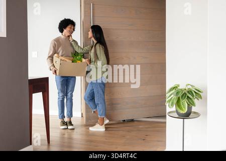Couple souriant diversifié déballage de l'épicerie fraîche dans l'entrée, avec boîte en carton et plante en pot Banque D'Images
