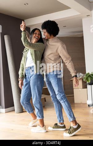 Danser un couple diversifié riant ensemble dans un salon moderne, avec des boîtes mobiles et un tapis roulé Banque D'Images